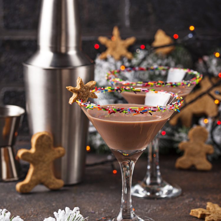 Zwei festliche Sugar Cookie Martinis mit braunem Getränk, bunten Streuseln am Glasrand und Sternenplätzchen, umgeben von Weihnachtsdekoration und einem Shaker.